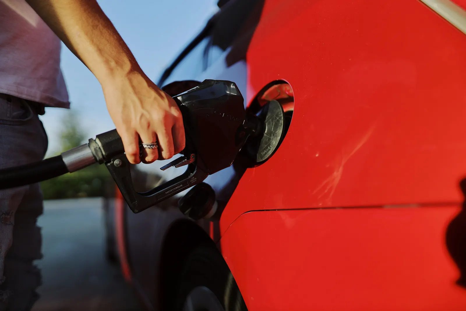 Mão segurando o bico da bomba enquanto abastece um carro vermelho sob o sol